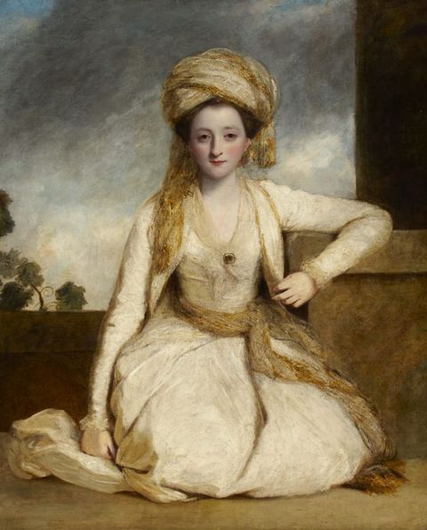 Painting. Portrait of a pale-skinned woman sitting on her knees in front of a ledge under a cloudy sky. She looks at us with hooded, dark eyes. She wears a gold-coloured turban and sash around her waist, over an ivory-white dress.