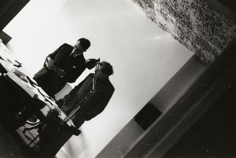 Black and white photograph. Two men wearing suits stand behind a desk, both looking at an open book one of them holds. The camera is tipped so their bodies angle from the lower left corner to the upper right.