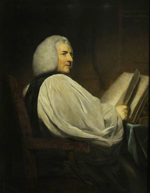 Painting. Shown from the lap up, a pale-skinned man sits with his body facing away from us to our right. He turns his head to look off to our right under dark brows. He has puffy white hair and a sharp nose. He holds a book open in front of him. The white sleeve we see covers most of his body.