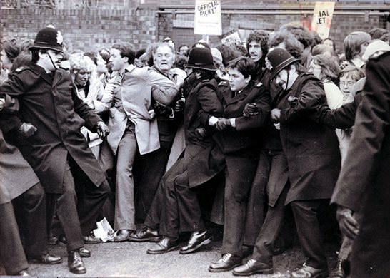 Black and white photograph. Uniformed police officers stand arm in arm with people in coats as crowds press from both sides. A sign held high reads, “Official Picket”.