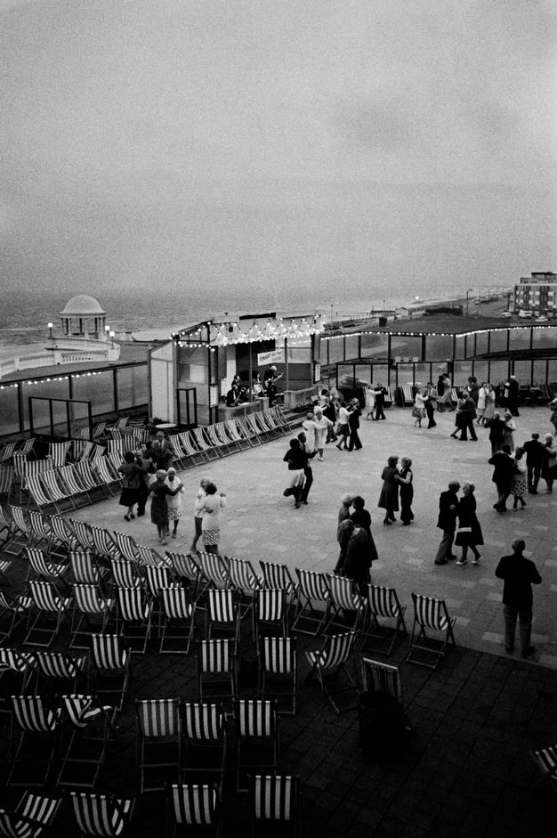 Black and white photograph. Couples dance on an open-air dance floor by the sea. Empty striped sling patio chairs are set up around the dance floor.