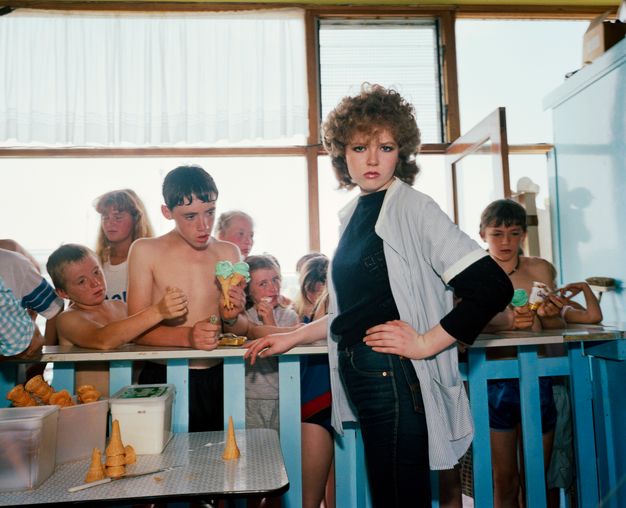 Colour photograph. A woman with hooded eyes under a poof of brown bangs looks at us, her downturned lips parted and her hand on her hip. She stands on our side of an ice cream counter, which is crowded with children on the other side.