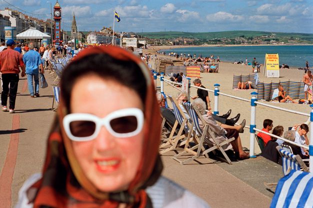 Colour photograph. The head and shoulders of a smiling, pale-skinned woman close to the camera are out of focus. The white frames of her sunglasses seem to glow. In sharper focus, a boardwalk crowded with people and a beach stretch into the distance beyond.