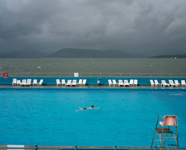 Colour photograph. A single person swims in a jewel-toned blue pool as slate-grey clouds approach across the smoky-green water of a bay. Empty white lounge chairs line a blue fence on the far side of the pool.