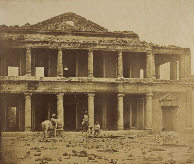 Sepia-toned photograph photograph. Four dark-skinned men and a white horse stand in front of the ruins of a two-storeyed, colonnaded structure. Rubble is strewn along the street in front of us.