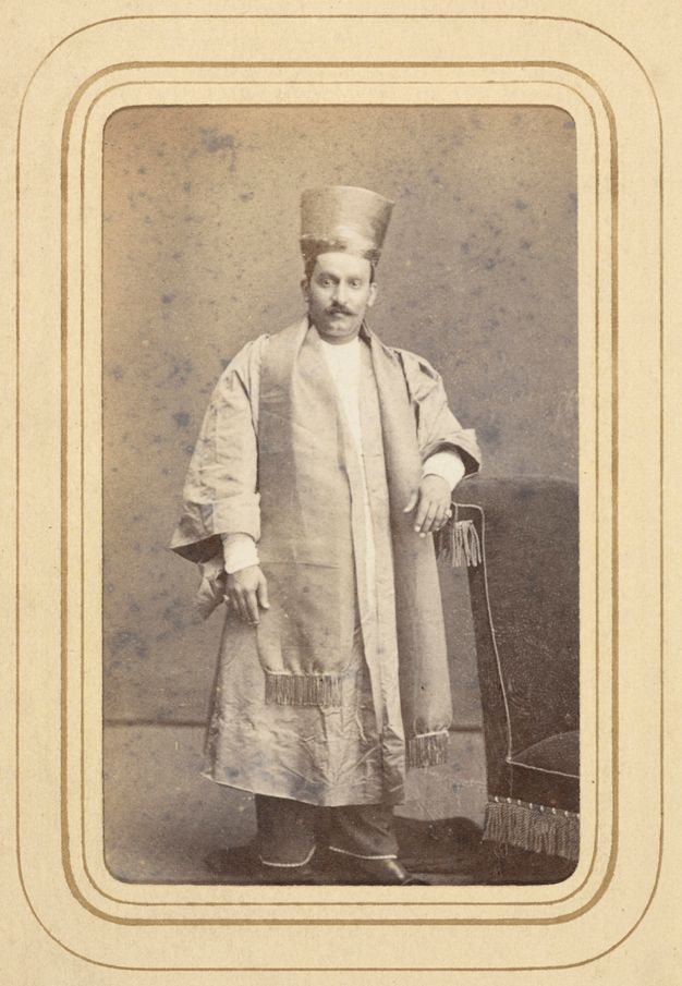 Sepia-toned photograph. Full length portrait of a man is set into a gold bordered carte-de-visite. He wears a tall, conical hat and a long scarf over an ankle-length robe.
