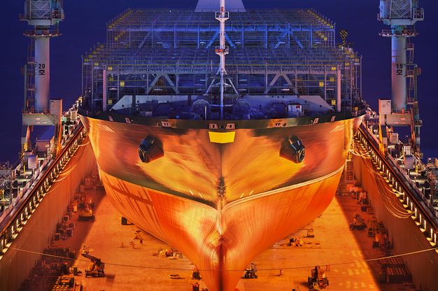 Colour photograph. The hull of a ship glows warm orange in a shipbuilding yard. The area beneath it, where machinery and supplies are stored, is also bathed in orange light. The scaffolding behind it and the space beyond, presumably water, are sapphire blue.