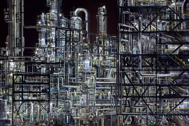 Colour photograph. Gleaming silver and jet-black pipes create an impenetrable web against a night sky. Lights among the equipment register as starbursts.