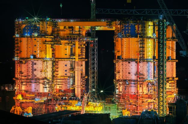 Colour photograph. The attached silo-like structures of an offshore plant are vivid orange, yellow, and blue against a velvet-black sky.