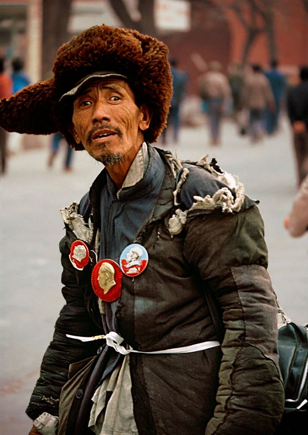 Man with medium brown skin looks at us on a city street. He wears a fuzzy brown hat with ear flaps and two winter coats tied around the waist with a white band. Three medallions or pins are affixed over his chest, each showing a person in profile. His eyebrows are gathered, and his hollow cheeks lined.