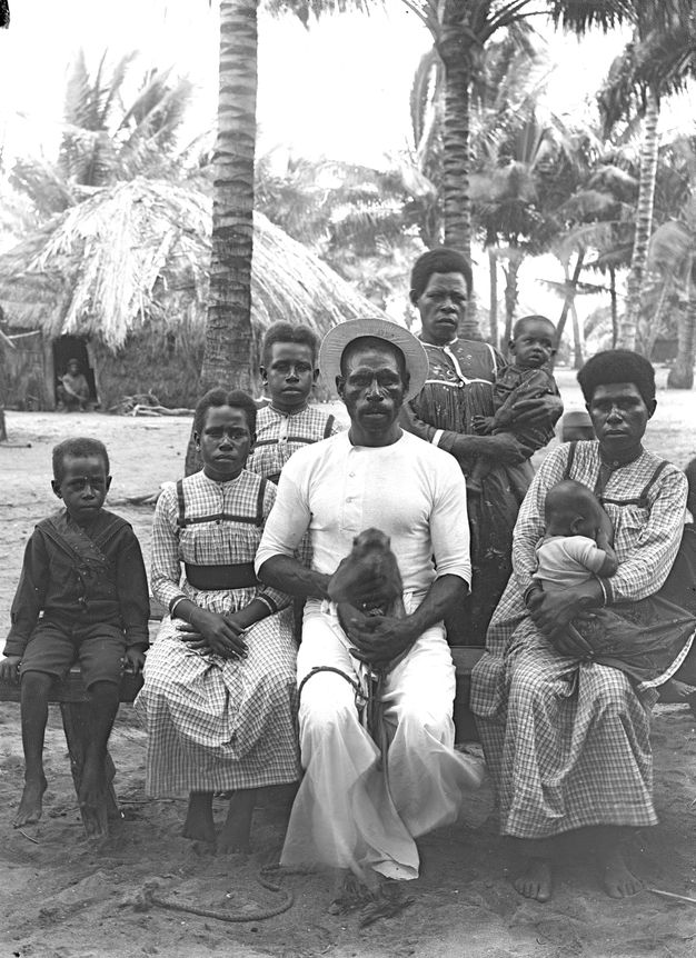 Black and white photograph. Eight men, women, children, and babies sit in front of a palm trees and a rounded, thatched structure. They all have dark skin and hair. Three of the women and girls wear dresses with the same checkered patterned. A man wearing white in the middle holds a monkey in his lap.