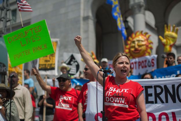 Colour photograph. A man and woman wearing red T-shirts reading “Shut. It. All. Down.” stand in front of a crowd holding signs. The woman raises a fist and speaks into a microphone. Farther back, the man holds a neon green sign reading “Disrupted Lives Porter Ranch”.