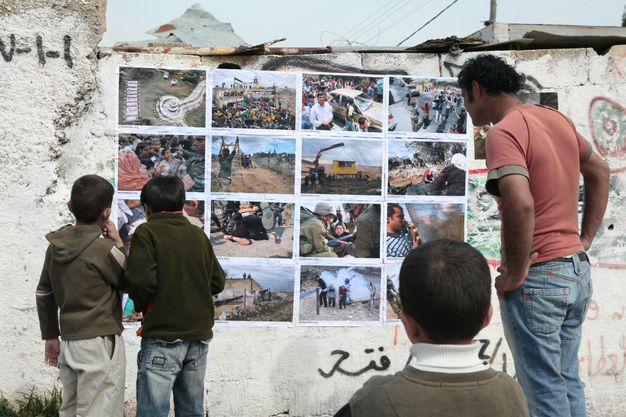 Colour photograph. Three children and a man look at a grid of sixteen colour photographs hung four by four on a graffitied wall. The posted images show mostly groups of people, including a clash of military personnel and civilians, a person trampled underfoot, and a smoke bomb.