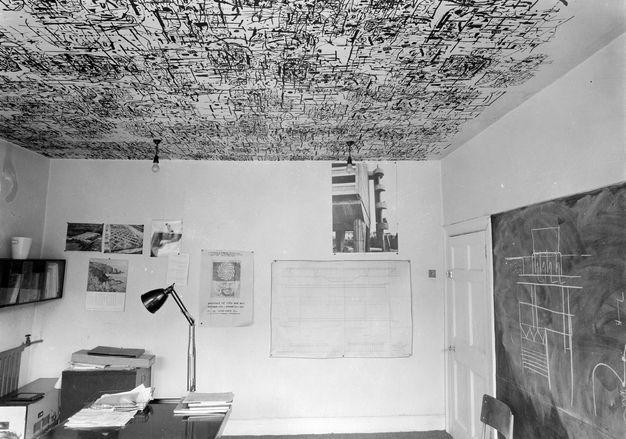 Black and white photograph. The ceiling of an office is decorated nearly edge to edge with an abstract design of black lines. Works of art hang on the walls below.