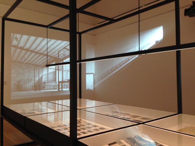 Installation photograph. Black and white film projects over glass cases holding printed material and perhaps photographs in the corner of a gallery. One film still shows an empty room. The other looks like the projector is so close to the wall that the image is distorted, becoming a ray of light along one wall and a sliver of film on the other.