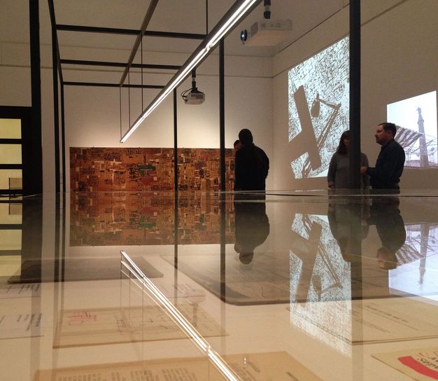 Installation photograph. We look across the top of a glass case holding documents at film projections and a work of art, mostly in tones of brown with a few touches of green and red, hanging across from us. The artworks and the few people who stand in the exhibition are reflected in the top of the glass case.