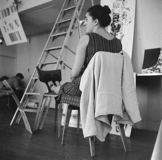 Black and white photograph. From close to the ground, we look up at a woman sitting in one chair with her feet propped up on another. She looks to our left in profile in a gallery with artwork being installed.