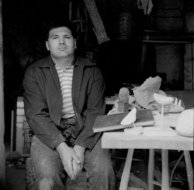 Black and white photograph. A man sits looking at or towards us from next to a table holding a book and small pieces of sculpture. His hands are together between his knees, and his chin is lifted.