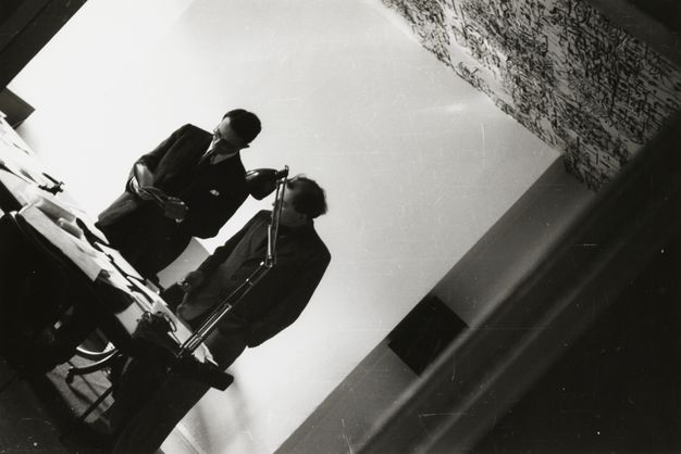 Black and white photograph. Two men wearing suits stand behind a desk, both looking at an open book one of them holds. The camera is tipped so their bodies angle from the lower left corner to the upper right.