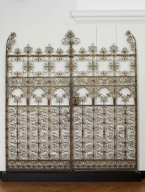 Gates. Gilded wrought iron gates curl and swirl in an intricate pattern reminiscent of filigree. The shadows the gate casts on the white wall behind it make it appear blurry, like double vision.