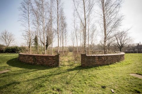 Colour photograph. A circular brick wall on a grassy field encloses spindly trees. A pass-through on either side is well trodden.