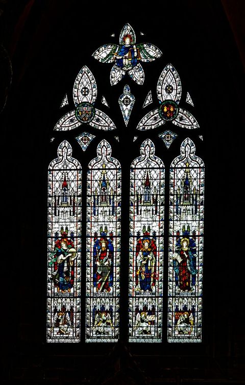 Stained glass window. Four people stand, each in a tall narrow pane, and each holds a musical instrument. The four panels are topped with trefoil and five-lobed windows. Flecks and slivers of glass between the larger components glint like sparks against the black surround.