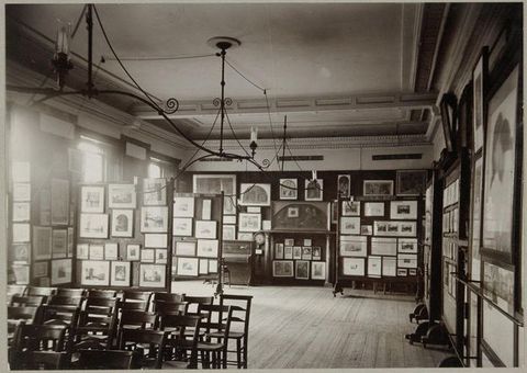 Black and white photograph. Works of art hang from floor to ceiling on every wall of this room. Wooden chairs are set up in rows in the lower left corner of the composition, facing something behind us and to our left.