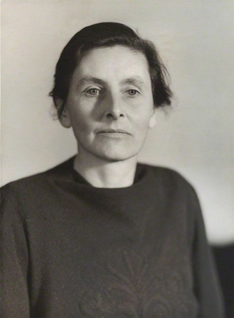 Black and white photograph. Shown from the chest up, a woman faces us but looks down and to our right. Her dark hair is pulled back, and she has a strong, straight nose and wide mouth in her square-jawed face.
