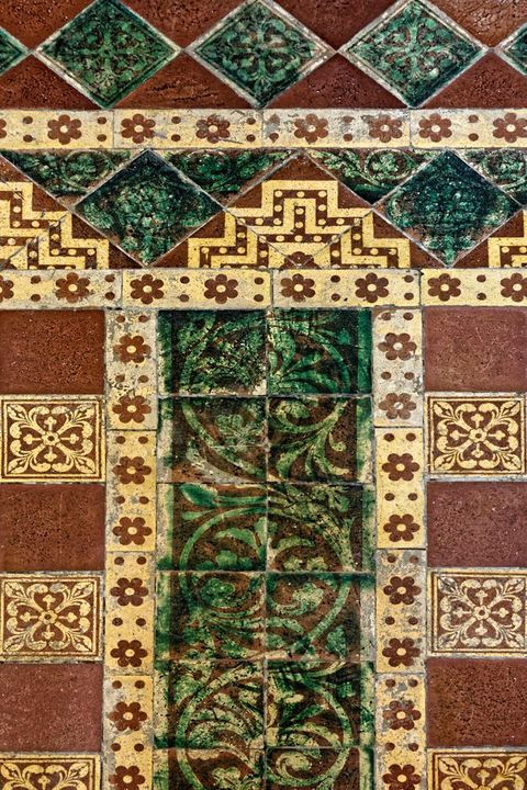 Pavement. A detail shows forest-green tiles decorated with curling vines, set into a framework of longer, thin tiles with rosettes against a cream-white background. Other tiles, in dark red or floral-patterned, create an intricate design.