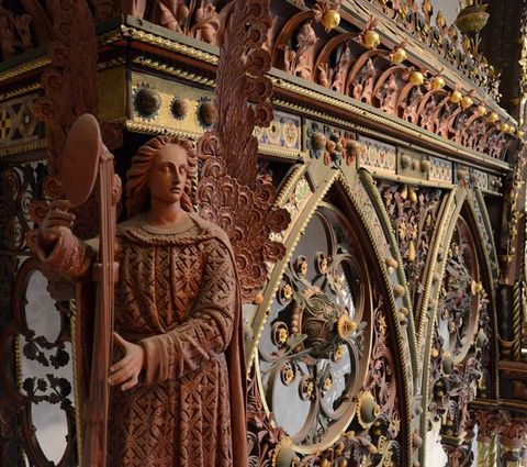 Choir screen. Colour photograph with detail view of a muted red winged angel holding a large standing instrument against intricately painted tracery.