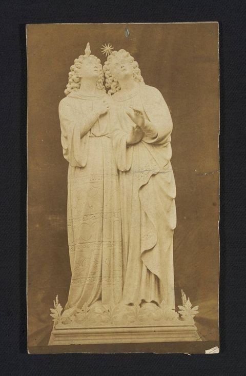 Sculpture. Printed in sepia-brown tones, this photograph shows a pair of statues sharing a base. They both have shoulder-length curly hair, wear long robes, and look up. One has a flickering flame over the head and the other a starburst.