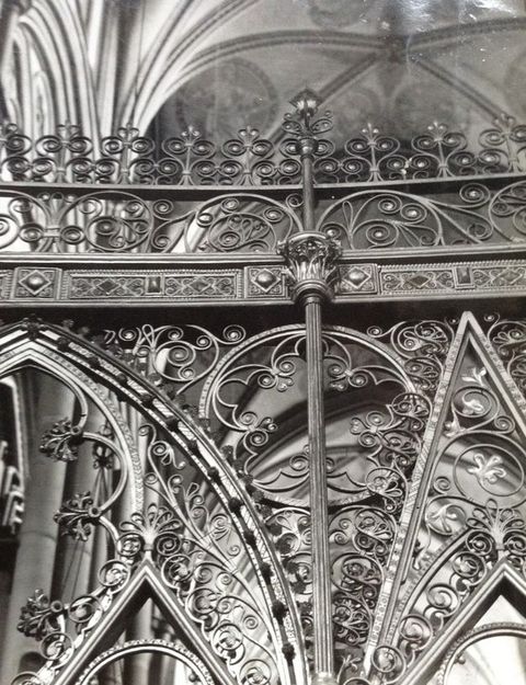 Choir screen. This black and white photograph focuses on the intricately swirling ironwork along the top edge of a choir screen. The vaulted ceiling of the church is glimpsed above.