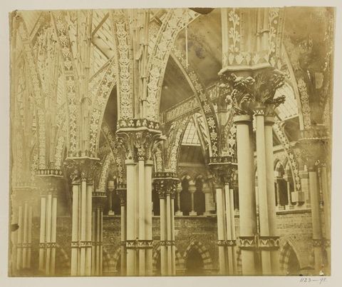 Architecture. Printed in sepia-brown tones, this photograph shows columns holding up heavily ornamented capitals and arches. Additional pointed arches are set into brick walls in the space beyond.