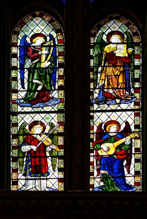 Stained glass window. Two pointed arched windows each show two winged angels playing musical instruments, one over the other, for a total of four. The clothing and objects are created with lapis-blue, emerald-green, crimson-red, and butterscotch yellow glass.