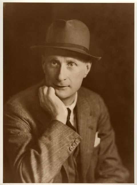 Sepia-toned photograph. A man shown from the waist up looks off to our left, his chin in one hand. He wears a pinstriped suit and a fedora hat. A white pocket square peeks up from behind the far lapel.
