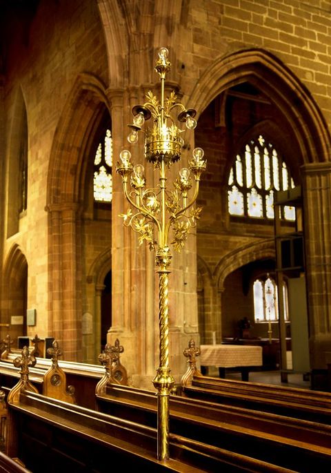 Metalwork. Installed so it rises from the back of a pew, a shiny gold light has two sets of electric bulbs on a tall, twisted shaft. Arms holding the naked bulbs reach out and up like a chandelier, to meet a few more arms bowing down around a crown-like form.