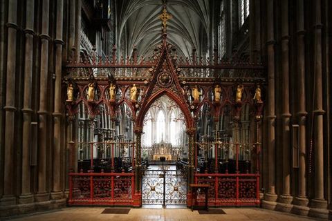 Choir screen. Colour photograph in which the delicate tracery of a red and gold screen is backlit from the bright transept beyond. Gold figures line the upper level of the screen. The central opening is a gated archway.