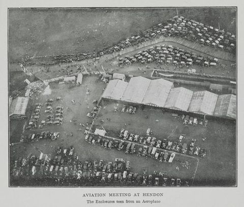 Black and white photograph. A high, aerial view of flat grounds. Six wide hangars are lined up near the centre of the composition. Cars are parked around the edges of the fields, and people dot the landscape. Text below reads, “Aviation Meeting at Hendon The Enclosures seen from an Aeroplane”.