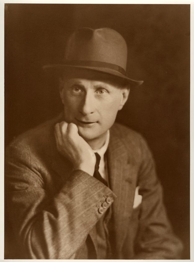 Sepia-toned photograph. A man shown from the waist up looks off to our left, his chin in one hand. He wears a pinstriped suit and a fedora hat. A white pocket square peeks up from behind the far lapel.