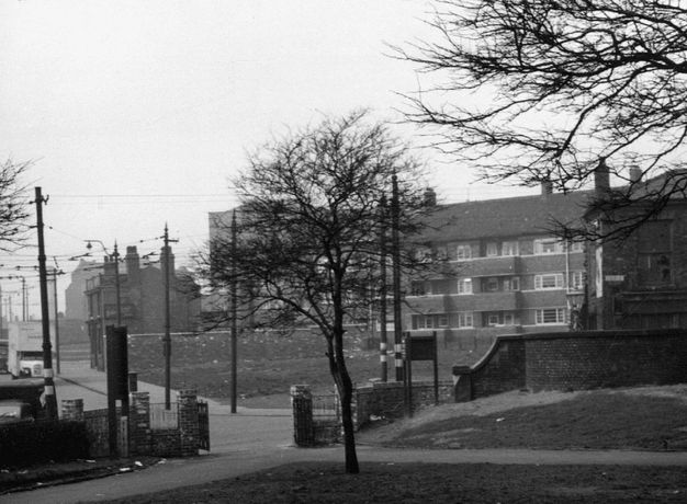 Black and white photograph. Litter-strewn pavements and lawns lead back to a block of flats. A moving van is parked in the street to our left, and utility poles and wiring create a web across the picture.