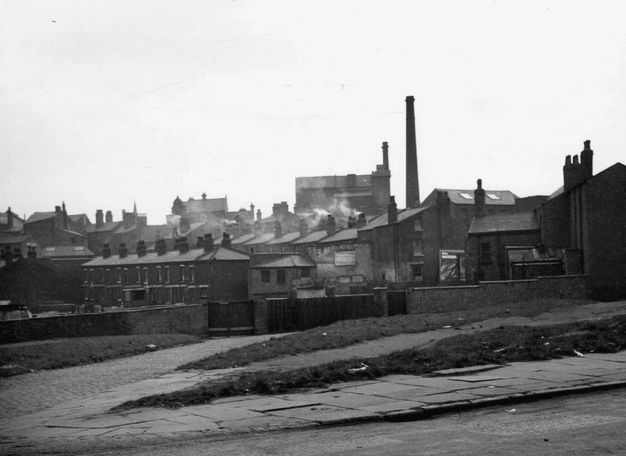 Black and white photograph. A street slopes sharply downhill and meets a stone-paved road at a hairpin junction in the lower left corner of this composition. Rows of buildings puffing smoke from chimneys fill the middle distance.