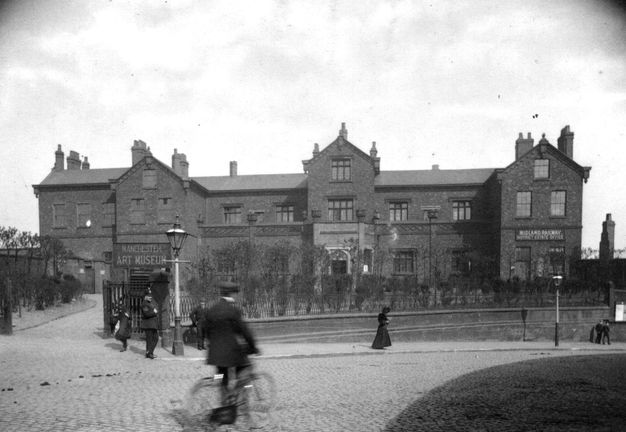 Black and white photograph. A road curves in front of a long brick building with three peaks over projecting bays spaced along its length. A sign over a door on the leftmost bay reads, “Manchester Art Museum”. A sign over the rightmost reads, “Midland Railway District Centre Office”.