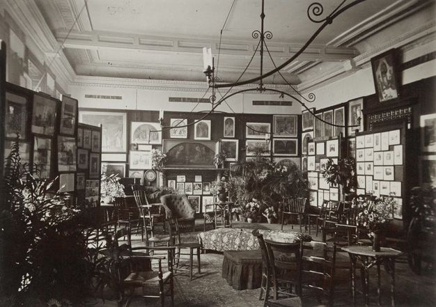 Black and white photograph. The walls of a room are hung densely with paintings and works of art. About a dozen chairs, sofas, and small tables are set up in the space. Sprays of potted plants add to the cluttered feeling.