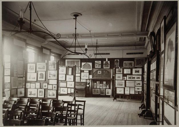 Black and white photograph. Works of art hang from floor to ceiling on every wall of this room. Wooden chairs are set up in rows in the lower left corner of the composition, facing something behind us and to our left.
