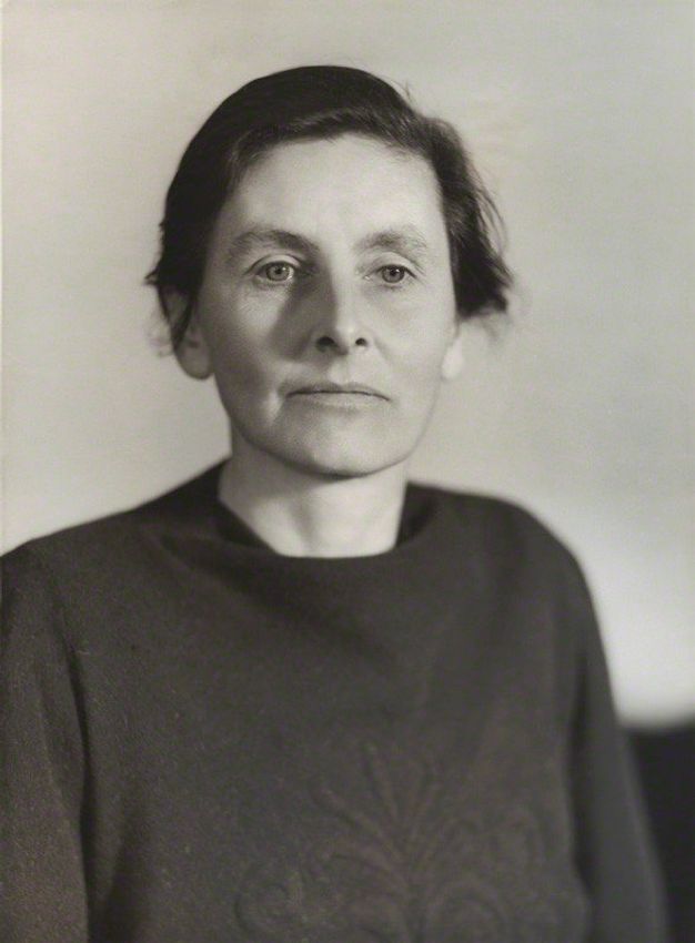 Black and white photograph. Shown from the chest up, a woman faces us but looks down and to our right. Her dark hair is pulled back, and she has a strong, straight nose and wide mouth in her square-jawed face.