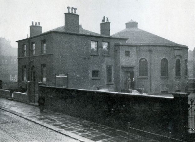 Black and white photograph. A blocky building with at least three chimneys is attached to a round structure like an oversized turret. A wall separating us from the structure is half as high again as a person walking on the pavement next to it.