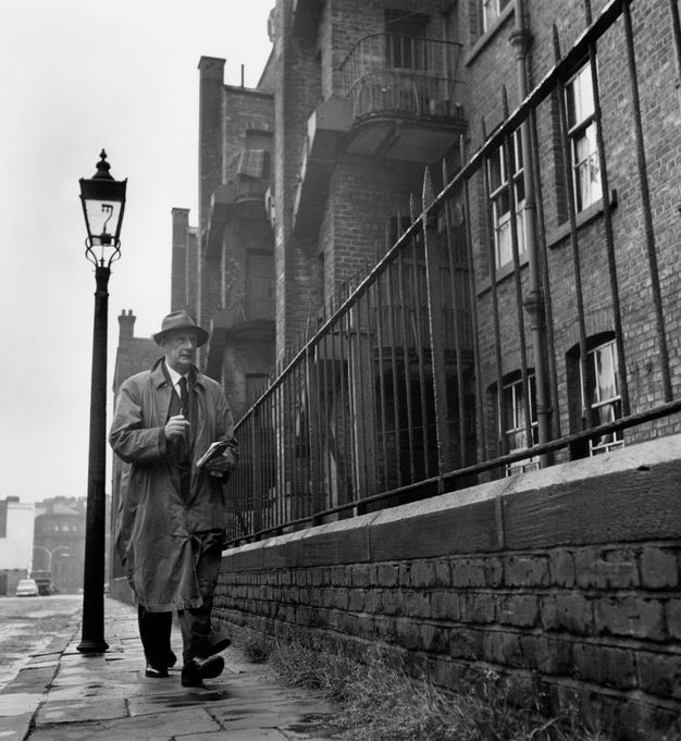 Black and white photograph. From low to the pavement, we look up at a man wearing a trench coat, walking towards us. To our left is a streetlight, and to our right, an iron fence and a row of brick buildings come at us at sharp angles.