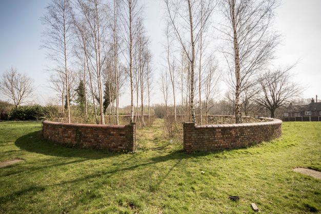 Colour photograph. A circular brick wall on a grassy field encloses spindly trees. A pass-through on either side is well trodden.