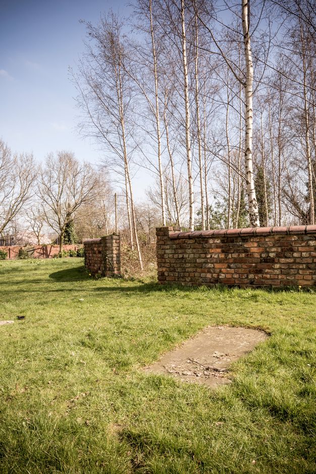 Colour photograph. An opening in a rounded brick wall leads to a copse of spindly trees. A rectangular patch of dirt mars the grassy field that surrounds the wall.
