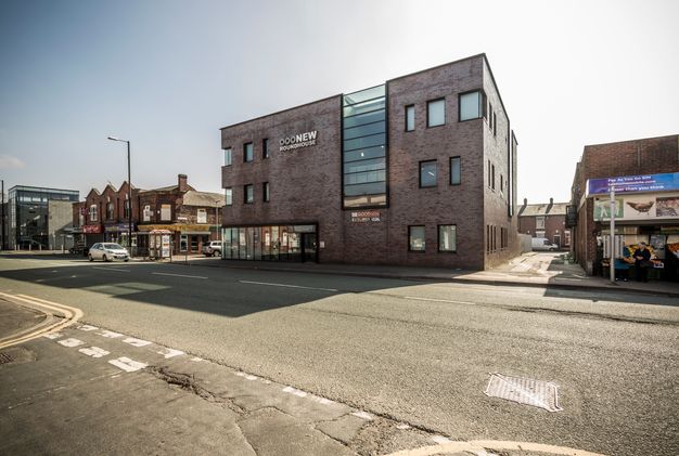 Colour photograph. Across a street from us, a blocky brick building is flanked by shops. Banks of windows cut down and across the façade facing us. A sign on that wall reads, “New Roundhouse”.